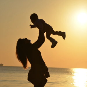 Woman hoisting her baby into the air, with the shoreline and setting sun behind them